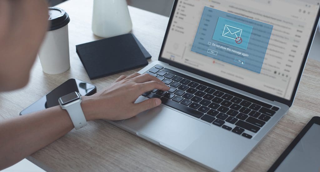 A person deletes and blocks a spam email on their laptop. A cup of coffee and a phone are in the background.