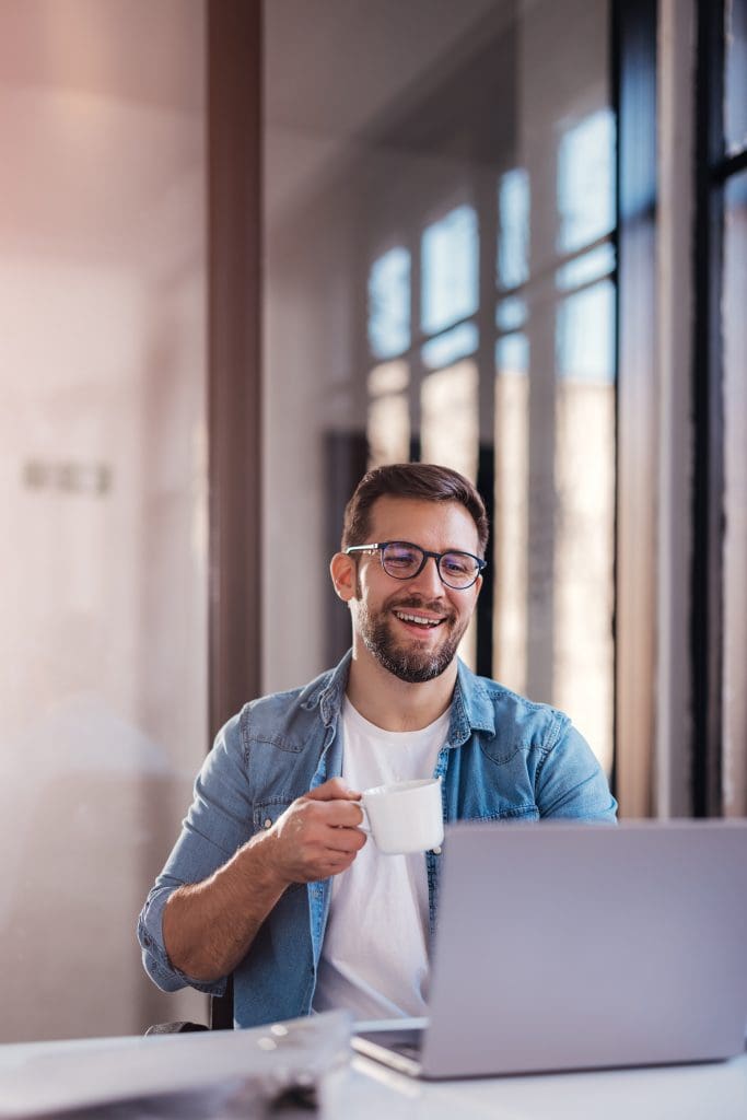 young man sitting by window in office, drinking coffee while working on laptop